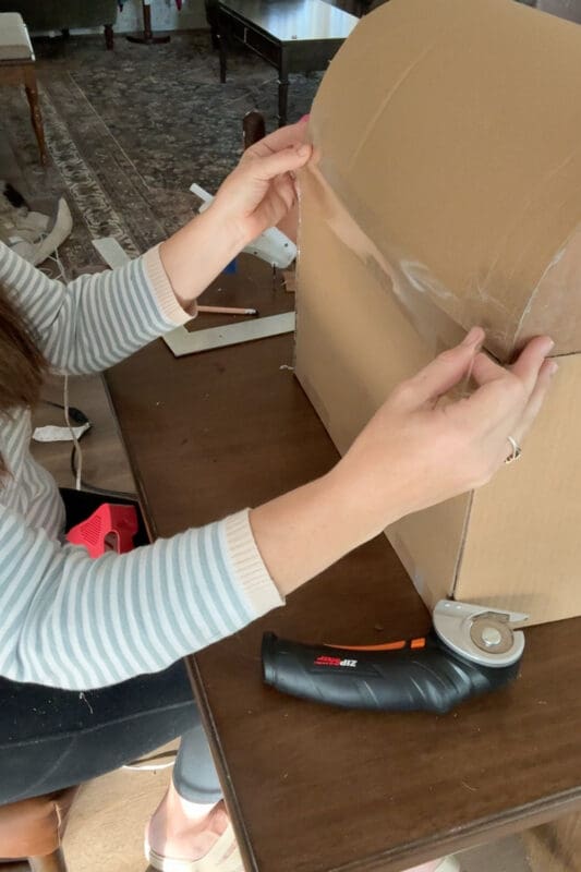 Crystel pressing clear packing tape along the back seam where the curved cardboard lid meets the box, creating an outside hinge for a DIY treasure chest, with a cordless electric cutter on the table beside it.