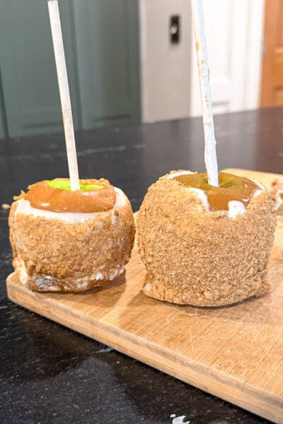 Homemade caramel apples with graham cracker crumb coating lined up on a bamboo cutting board on a black kitchen counter