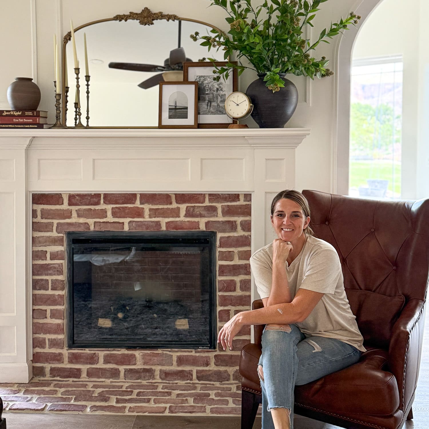 Crystel sitting in a brown leather wingback chair beside her finished DIY electric fireplace with a red brick surround, brick hearth, and white paneled mantel styled with a gold arched mirror and greenery