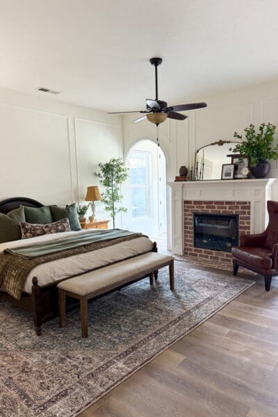 Bedroom makeover showing large bedroom ideas like picture frame molding, an arched doorway, and a brick fireplace with a gold arched mirror on the mantel, beside a leather wingback chair