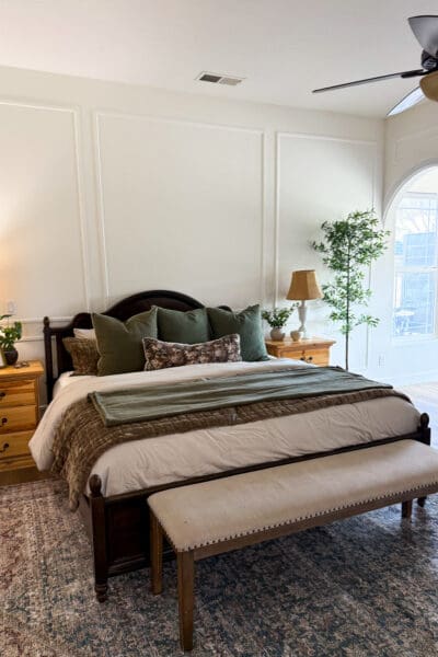 Dark wood bed with green and floral pillows, an olive velvet quilt, and a green throw set against a wall of picture frame molding, with a pine nightstand, a faux olive tree, and an arched doorway in a large bedroom