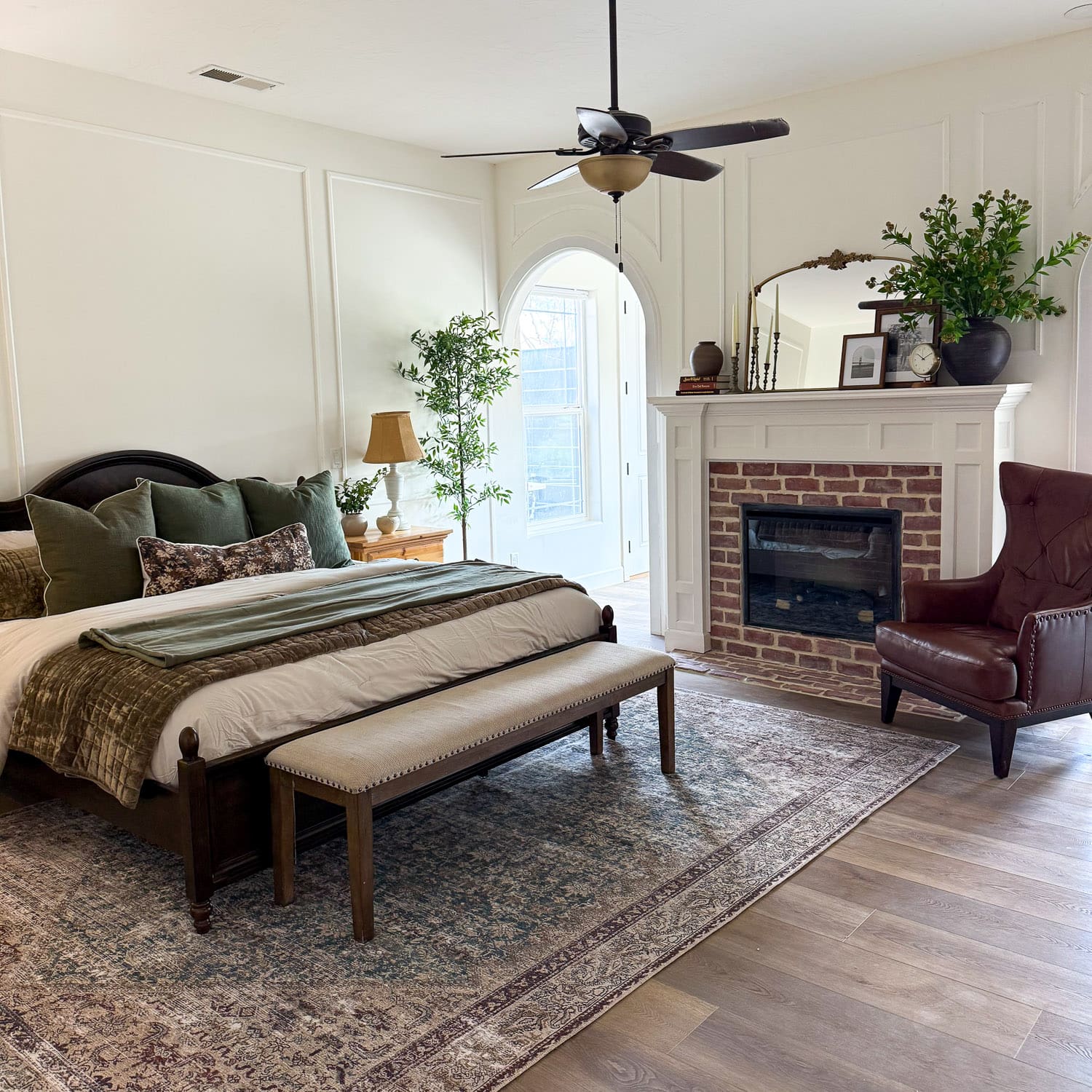 Large bedroom with a dark wood bed, green pillows, and an upholstered bench on a vintage style rug, facing a white mantel fireplace with a red brick surround, with picture frame molding on the walls and an arched doorway