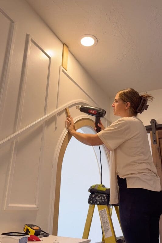 Crystel bending heated PVC molding to follow the shape of an arched doorway, holding a spacer against the wall to make curved picture frame molding in a large bedroom