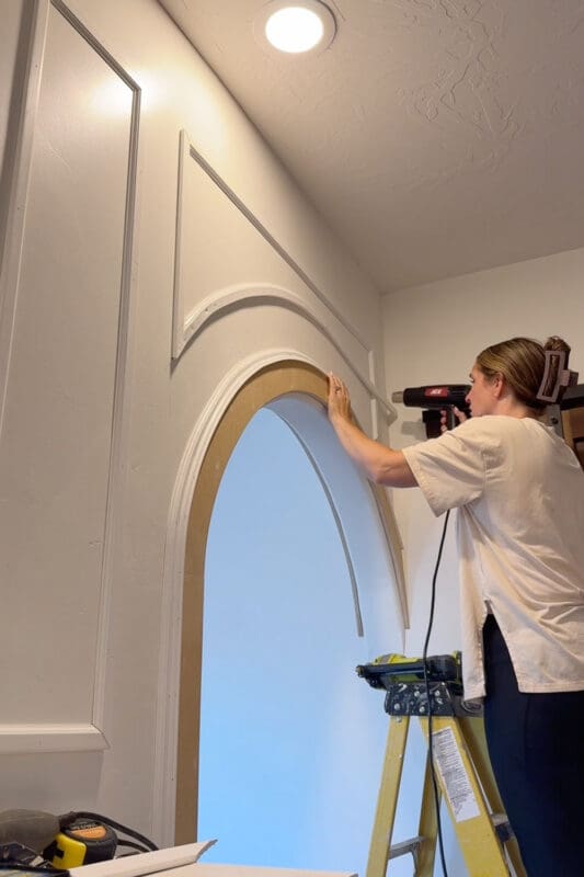 Finished curved picture frame molding box nailed above an arched doorway, while Crystel uses a heat gun to shape another piece of PVC trim along the arch