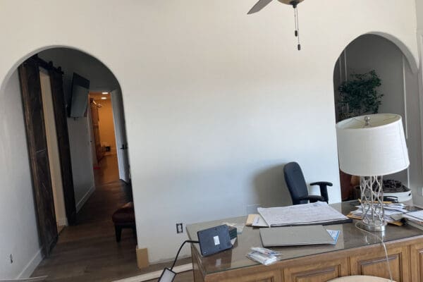 Back of the room divider wall between two arched doorways, with an office chair against the plain white wall, barn doors visible through one arch and a faux tree and picture frame molding through the other