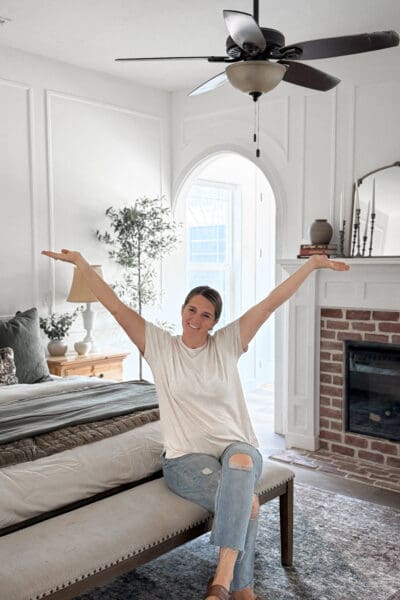 Crystel smiling with her arms raised while sitting on an upholstered bench at the foot of the bed, with the finished arched doorway, picture frame molding, and brick fireplace behind her