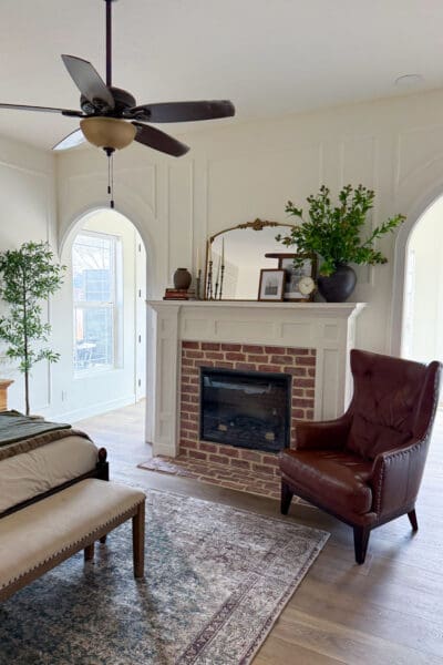 White fireplace mantel with a red brick surround and hearth, styled with a gold arched mirror, candlesticks, books, framed photos, a clock, and a vase of greenery, beside a leather wingback chair between two arched doorways