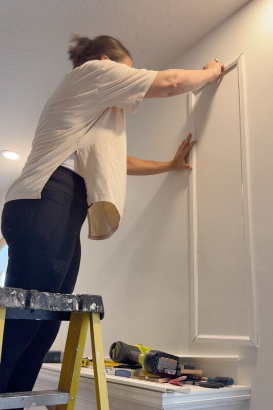 Crystel standing on a yellow stepladder pressing a PVC trim piece against the wall above the fireplace to build picture frame molding, with a brad nailer and tools resting on the mantel