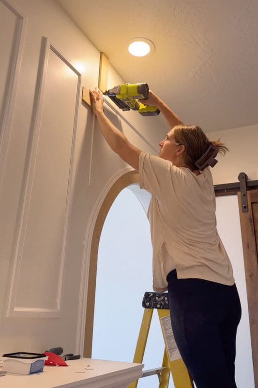 Crystel on a stepladder using a brad nailer to attach the top rail of a molding box above an arched doorway, with scrap wood spacers holding the gap from the ceiling and the neighboring box