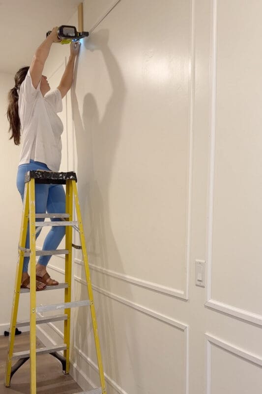 Crystel on a tall yellow ladder using a brad nailer to attach a molding rail near the ceiling, with a wood spacer set against the ceiling and two tiers of picture frame molding on the wall below in a large bedroom