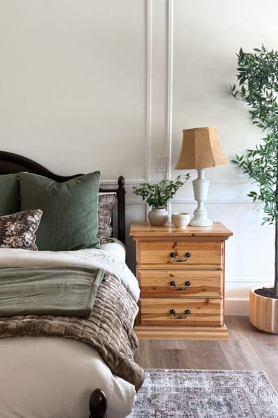 Pine three-drawer nightstand styled with a white lamp, a tan shade, a small vase of greenery, and a cream bowl beside the bed’s green pillows and velvet quilt, with a faux tree in a ribbed planter