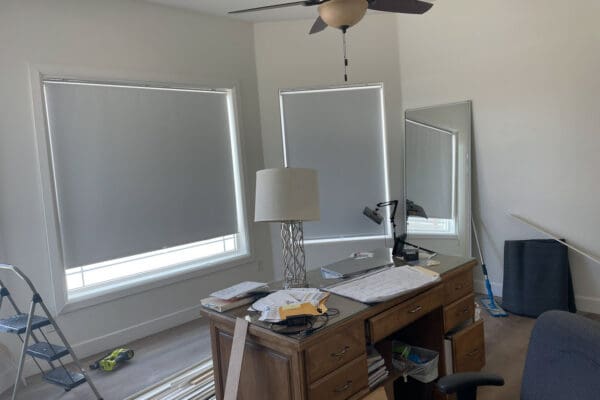 Back half of a large bedroom set up as an office in progress, with a wood desk covered in papers, a silver table lamp, two large windows with gray roller shades, a leaning floor mirror, and trim pieces on the floor