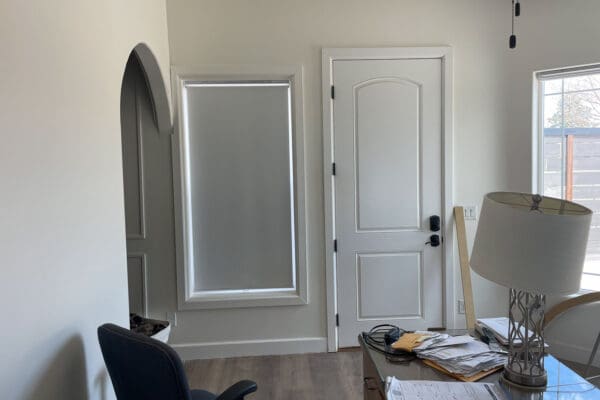 Office side of the divided bedroom showing a tall window with a gray roller shade next to a white exterior door, with an arched doorway on the left and a desk lamp in the foreground