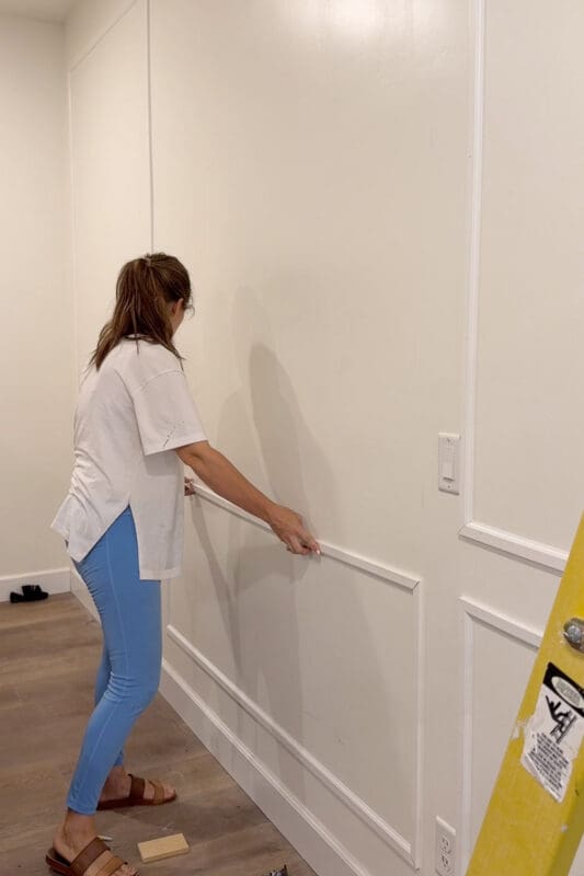 Crystel pressing the top rail of a lower molding box onto the bedroom wall where the bed will go, with taller picture frame molding boxes above and a scrap wood spacer on the floor